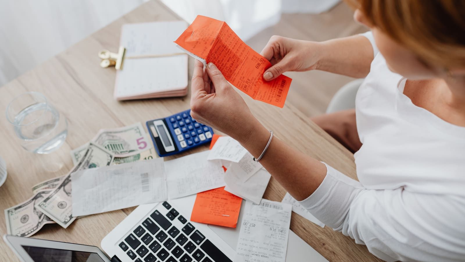 A woman sorts receipts next to a calculator, cash and a laptop on her desk