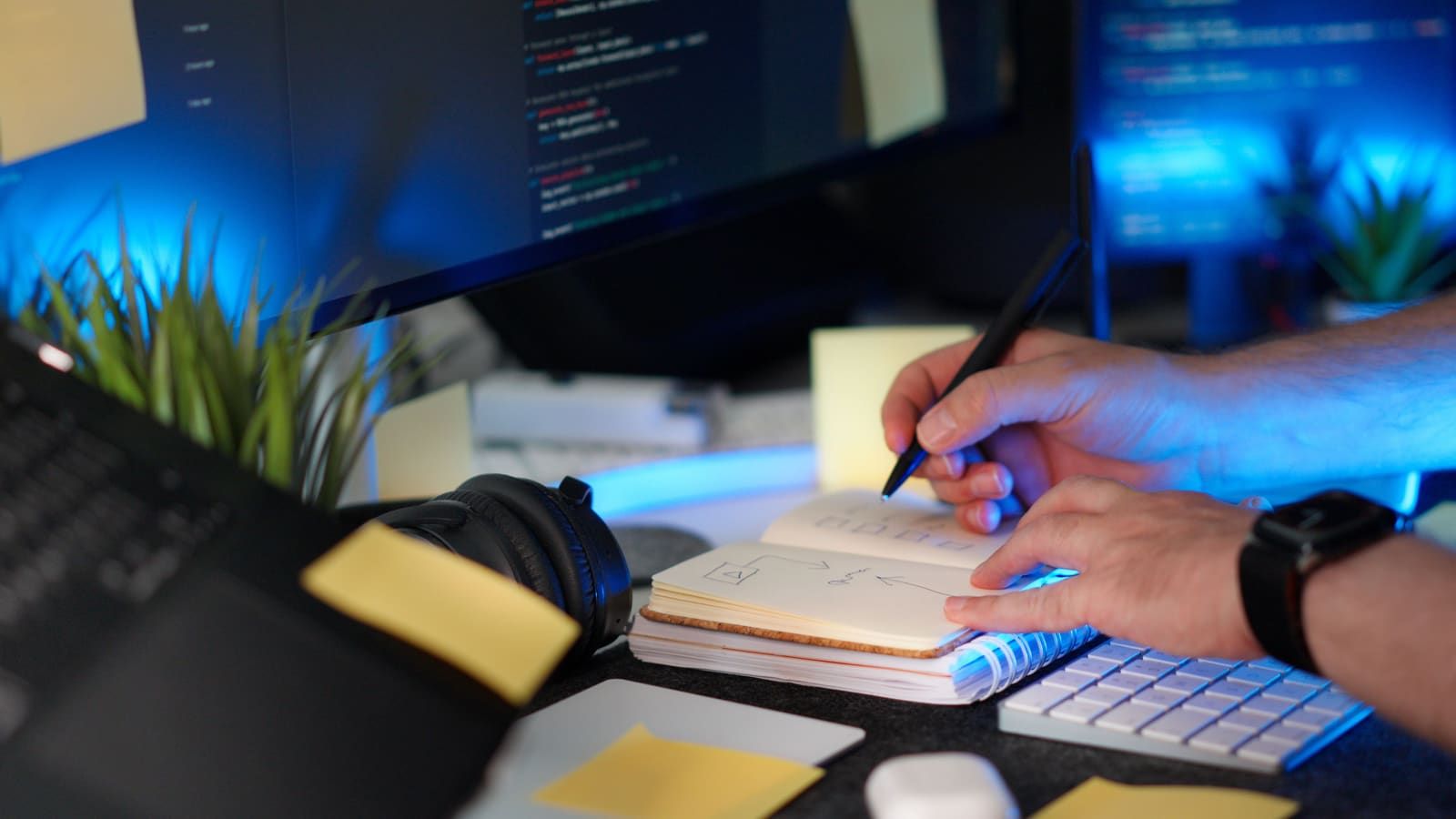 A developer writes notes in a notebook beside a keyboard, with code on the monitor