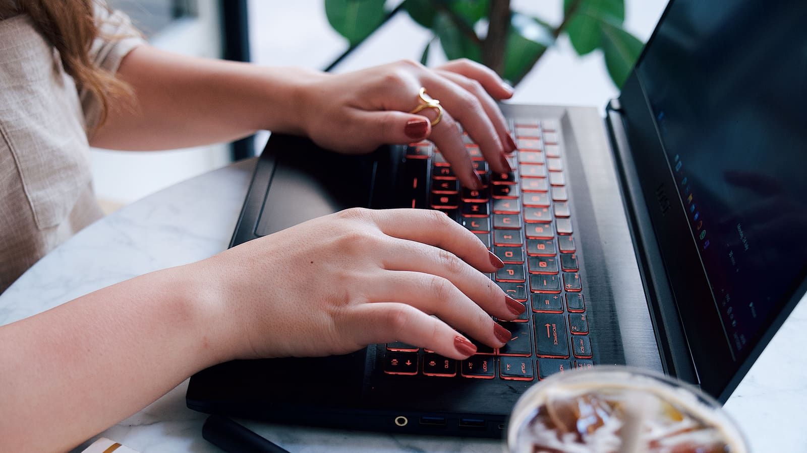 A young woman typing on a laptop at a café table with coffee and a notebook nearby
