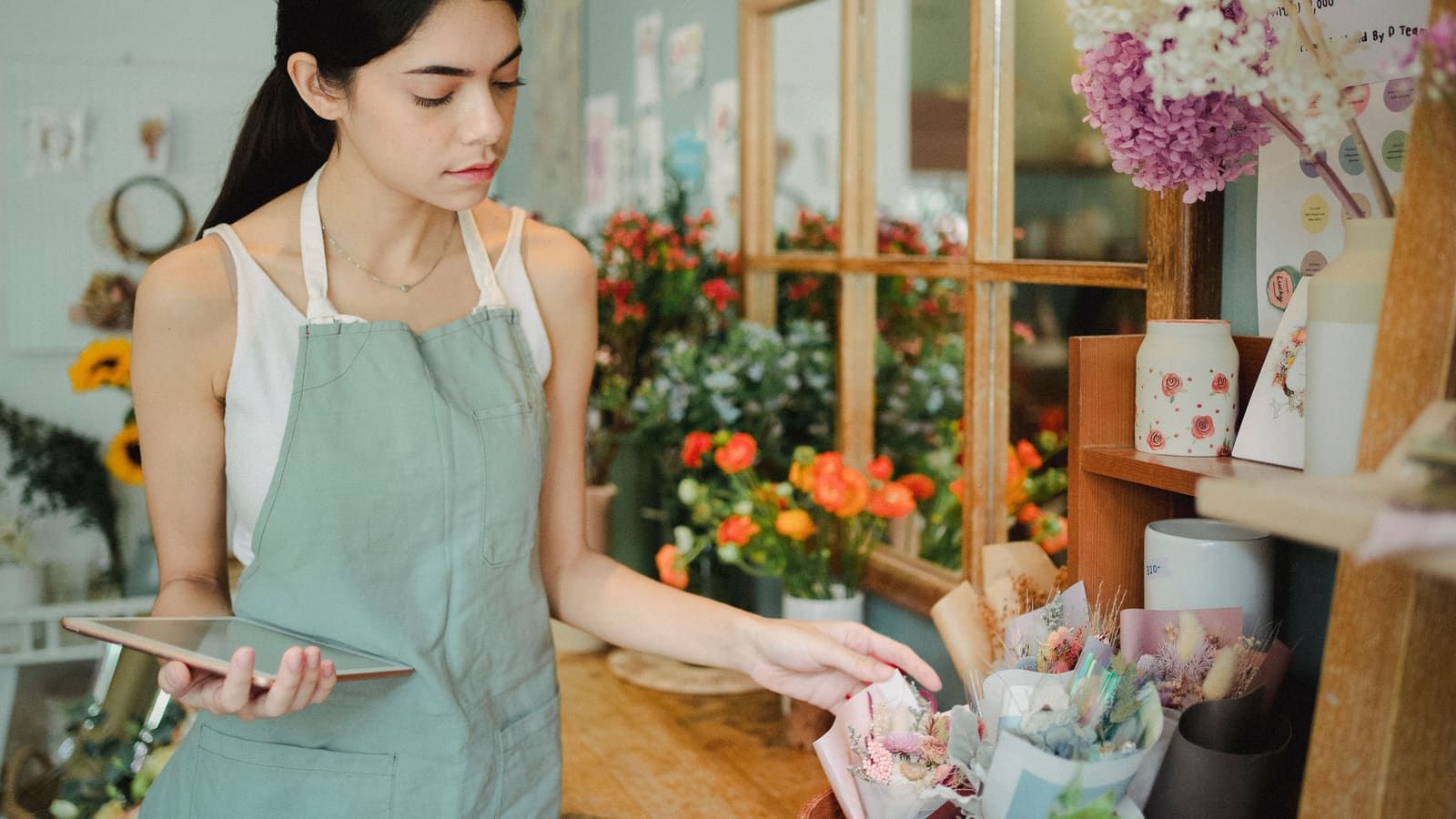 Concentrated female florist with dark hair standing in floral shop with tablet near flowers in wrapper while checking order during work