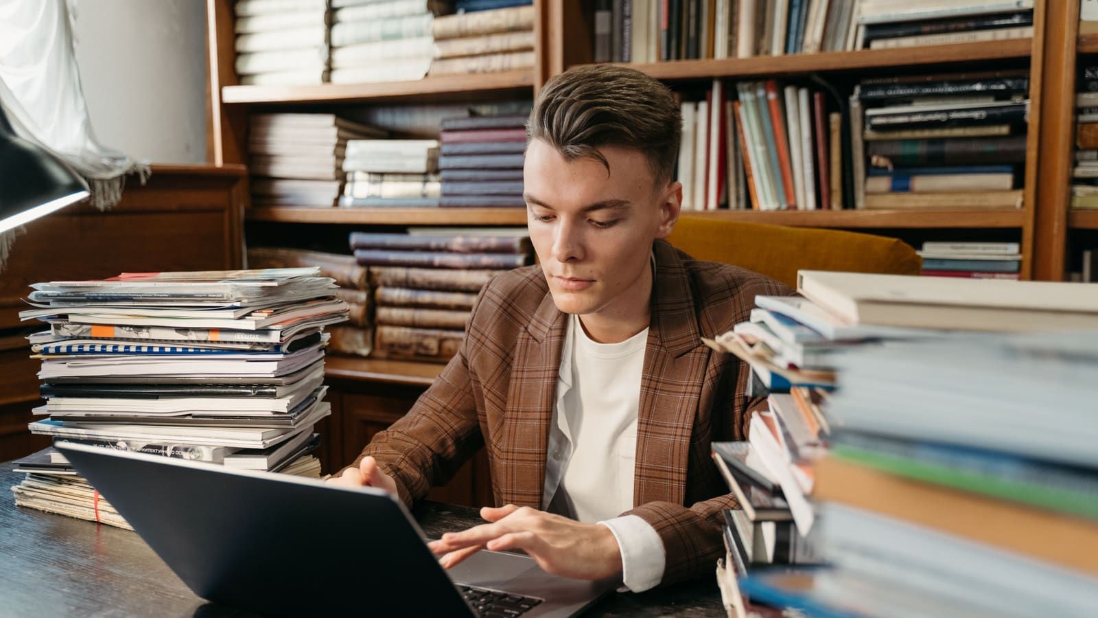 A focused young man studies on a laptop amidst stacks of books in a cozy library setting