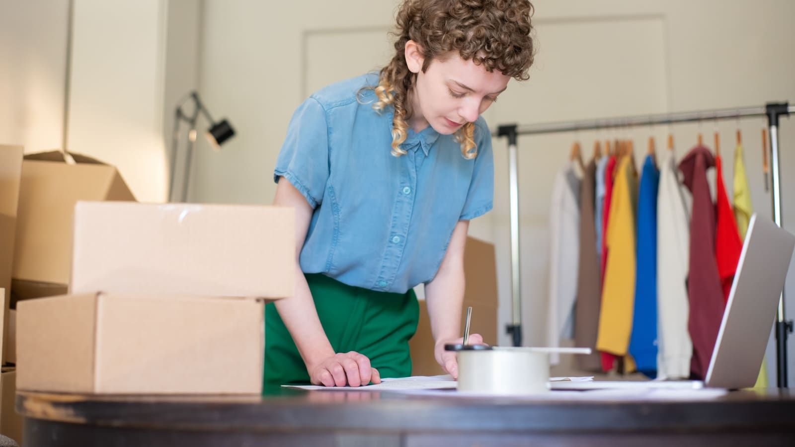 An online shop owner writes up orders among shipping boxes and a clothing rack
