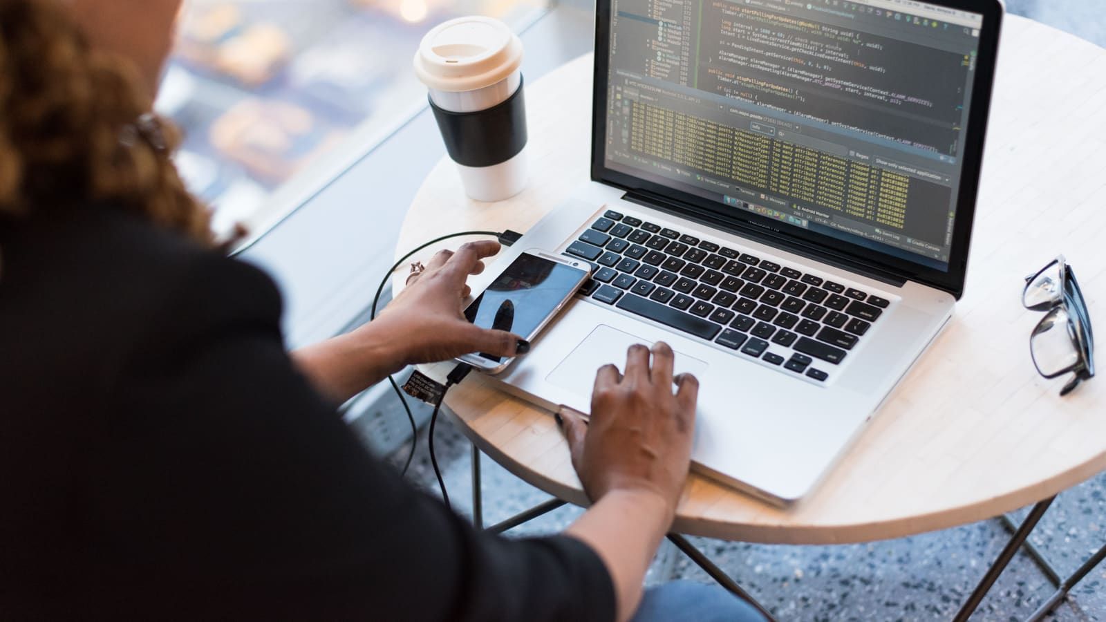 A developer checks her phone while working on code on a laptop at a café table